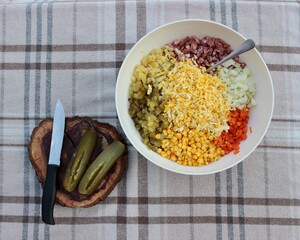 on a checkered tablecloth, a round bowl with chopped salad ingredients and a wooden board with a knife and a pickle..