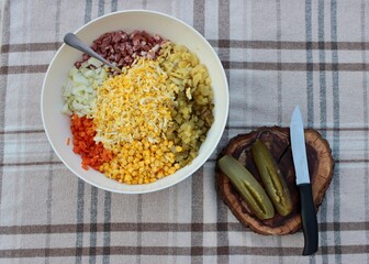 on a checkered tablecloth, a round bowl with chopped salad ingredients and a wooden board with a knife and a pickle..