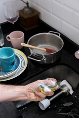 dirty kitchen utensils and person's hand holding a scourer with white foam
