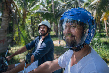 Two men on motorcycles with helmets in palm trees on a tropical island