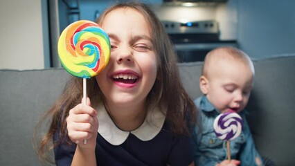 Kids With Lollipop. Two Happy Cheerful Children Eat Candy on a Stick at Home on the Couch. Baby Boy and His Older Sister Have Fun at Home Eating Candy