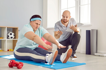 Fototapeta na wymiar Fat woman overcomes herself during hard sports workout with trainer. Instructor motivates and encourages overweight woman who is sitting on exercise mat trying to reach and touch her toes with hands