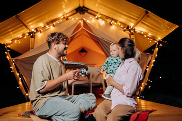 Happy family with lovely baby relaxing and spend time together in glamping on summer evening near cozy bonfire. Luxury camping tent for outdoor recreation and recreation. Lifestyle concept - 601148881