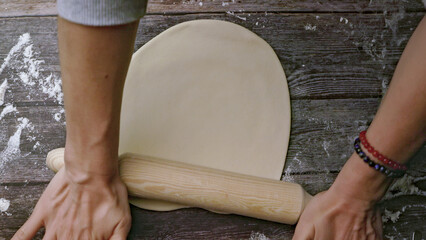 Top view: women's hands roll out the dough for a pizza base on a wooden table with a rolling pin.