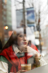 Pensive, beautiful woman in eyeglasses sitting in cafe, looking at window, holding drink