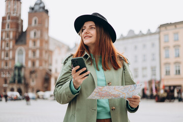 Fototapeta Beautiful stylish woman in hat walking on Market Square in Krakow on autumn day and holding mobile phone and map. Phone Communication. Urban lifestyle concept. Check social networks, booking hotel obraz