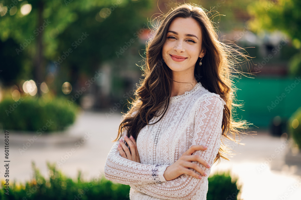 Wall mural Portrait of a beautiful young woman standing on a city street with arms crossed and smiling at the camera on a sunny summer day.