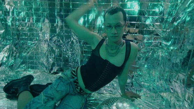 Young short-haired female model in trendy clothing sitting on the floor, posing with disco ball in studio with neon strobe light and foil backdrop. Creative portrait, shaky cam shot