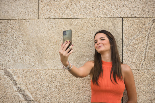 Young beautiful woman with straight brown hair and orange top, making a video call with her cell phone. Concept fashion, beauty, trend, mobile, smartphone, app, video, millennial.