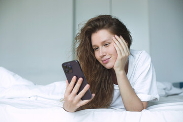 young happy woman use phone and lying in bed in bedroom in the morning, concept morning, phone addiction, social, habits