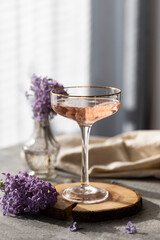 Pink cocktail in vintage glass with flower garnish on raw wood. Iced cold summer drink. Organic natural cocktail, lilac lemonade.  