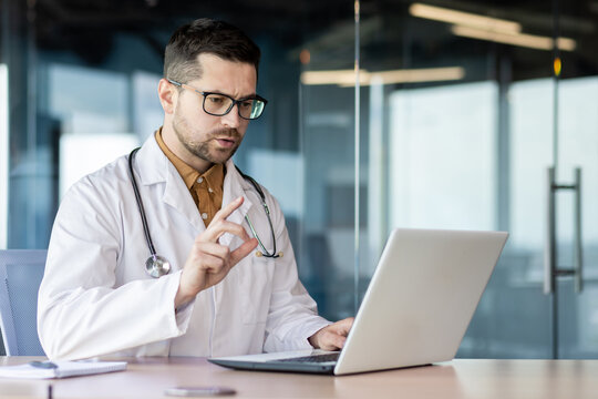 Young Serious Male Doctor Sits In The Office At The Table And Conducts Online Consultation, Remote Treatment, Video Call With The Patient. Discusses Therapy, Warns About Consequences