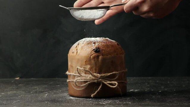Traditional Italian Christmas cake Panettone tied with a jute thread on the black background. Pastry Chef sprinkles powdered sugar through a sieve on the fruit Easter cake with raisins.