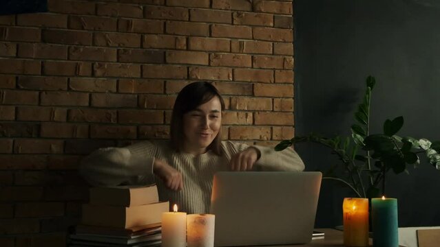 Happy caucasian woman using laptop at home during electricity outage or blackout with lit candles. Inspired woman raises her hands up after goal achievement. Success concept