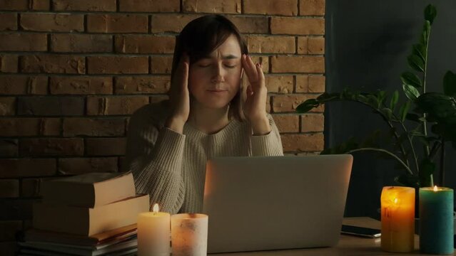 Caucasian woman is under stress using laptop at home during electricity outage or blackout with lit candles. Tired woman rubbing her temples and having a headache