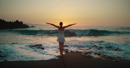 Fototapeta na wymiar Woman silhouette raises hands up on black sand beach at sunset. Twilight orange sky. Adult girl relax watching powerful sea waves landscape.