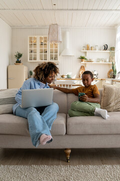 African American Mom Working On Laptop And Looking At Smartphone In Son's Hands Sitting On Couch At Home. Teen Boy Browsing In Smartphone, Watching Videos. Working Busy Mom, Child Gadget Addiction. 