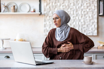 A young Arab woman in a hijab is working, studying at home using a laptop online, holding her chest, on the side of her body. feels pain, discomfort.