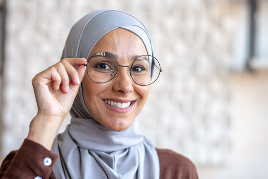 Close-up photo. Portrait of a young beautiful Muslim woman in a hijab smiling looking at the camera and adjusting her glasses with her hand.