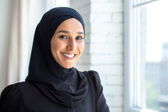 Close-up photo. Portrait of a young beautiful Muslim woman in a black hijab looking confidently and smiling at the camera, standing indoors by the window.