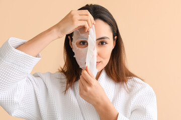 Young woman applying sheet mask on beige background, closeup