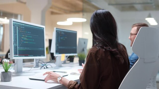 Side view of hardworking female programmer speaking with partner while working on computer. Program code visible on screen. Young people communicating in office. Developers concept.