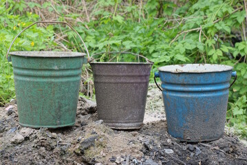 three old dirty metal enameled buckets stand on the gray earth among the green vegetation on the street