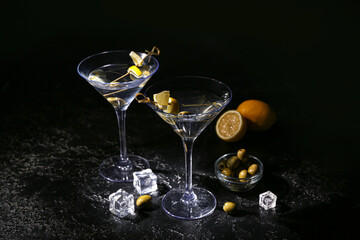 Glasses of tasty martini, ice cubes, green olives and lemon on dark background