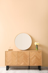 Stylish mirror and vase with narcissus flowers on wooden cabinet near beige wall