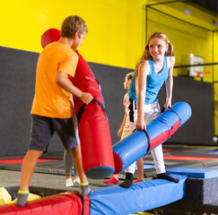 Happy child school boy and teenage girl friends fighting by inflatable logs while spending weekend together in entertainment center