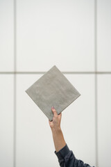 Female Hand Holding Square Gray Tile Against White Tiled Background