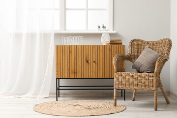 Stylish wooden cabinet and cozy wicker armchair near window