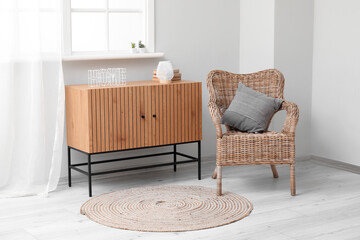 Stylish wooden cabinet and cozy wicker armchair near window