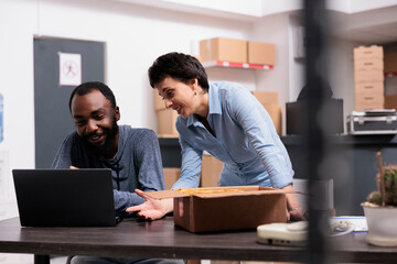 Workers finising packing trendy blouse while checking retail drop shipping package on laptop computer working in warehouse. Diverse team putting orders in cardboard boxes, distribution center concept