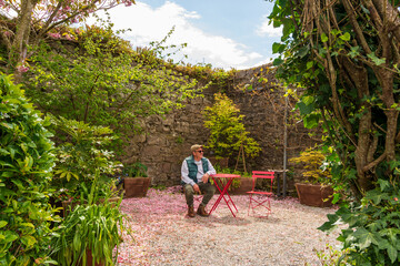 The image depicts a senior man sitting in a red chair in a garden surrounded by vegetation.