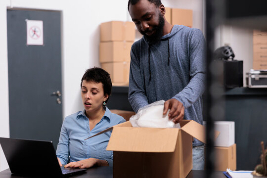 Storehouse worker putting client order in carton box wrapping with bubble wrap while supervisior woman checking transportation detalies on laptop computer. Warehouse job concept