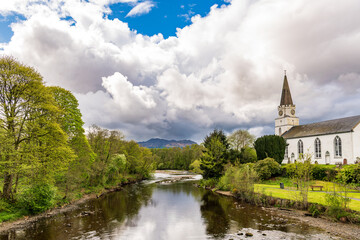 The river earn flowing past the white churh in the perthshire (scotland) village of comrie with green wooded banks, dramatic white clouds and reflections in the water.