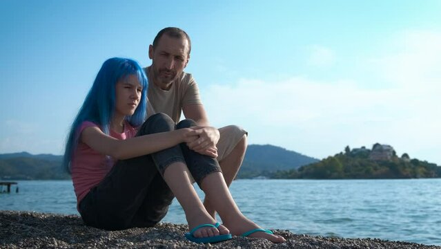 Parent talk by tropical beach. A father with her blue haired daughter pass time on the shore and speak about family trouble during summer vacation.