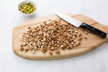 Wooden board with chopped hazelnuts and almonds and a bowl of peeled pistachios on a light gray background. Stage of cooking delicious homemade dessert