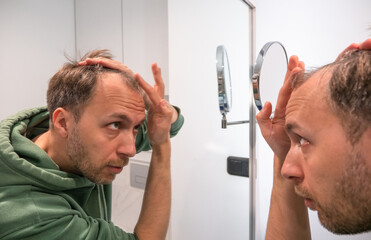 Balding middle age man looks in mirror and checks how much hair he lost. - obrazy, fototapety, plakaty