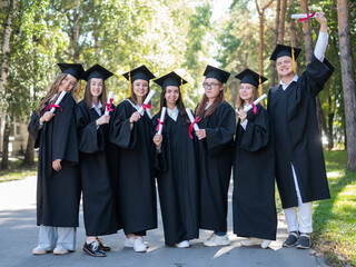 Row of young students in graduation gowns outdoors showing off their diplomas. 