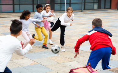 Cheerful tween schoolchildren gaily spending time together on warm spring day, playing with ball near school building.