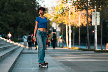 Fototapeta premium bella mujer adolescente estilo 80s 90s andando en skate en la ciudad, con maquillaje, anteojos y jeans. en un día soleado al atardecer.