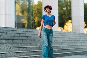 bella mujer adolescente estilo 80s 90s andando en skate en la ciudad, con maquillaje, anteojos y jeans. en un día soleado al atardecer. - obrazy, fototapety, plakaty