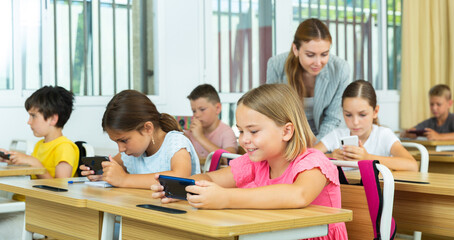 Schoolchildren sitting at desks in classroom. They're using their smartphones during lesson.