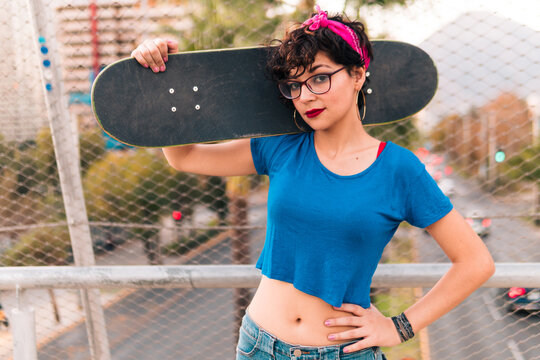 bella chica patinadora sonriendo y posando con su skate, con maquillaje, lentes y estilo 80s 90s en la ciudad.