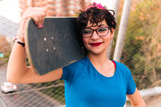 bella chica patinadora sonriendo y posando con su skate, con maquillaje, lentes y estilo 80s 90s en la ciudad.