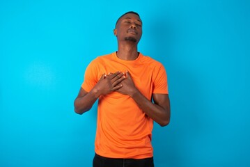 Handsome man wearing orange T-shirt over blue background smiling with hands on chest with closed...