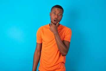 Handsome man wearing orange T-shirt over blue background Looking fascinated with disbelief, surprise and amazed expression with hands on chin