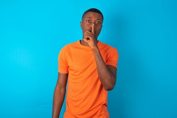 Handsome man wearing orange T-shirt over blue background makes silence gesture, keeps finger over lips. Silence and secret concept.